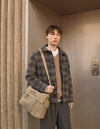 A.P.C. Men - Man wearing plaid wool jacket and grey wool pants holding a khaki green bag standing in front of a doorway.