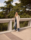 A.P.C. Women - Woman wearing cream colored coat and blue jeans on a bridge in the woods.