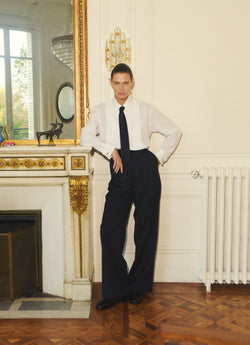 A.P.C. x Anastasia Barbieri campaign image, featuring a female model wearing a white shirt, a tie, dark pants, by a fire place