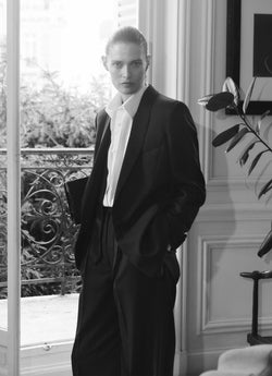 A.P.C. x Anastasia Barbieri campaign image, featuring a female model wearing a black suite and white shirt, standing by a window