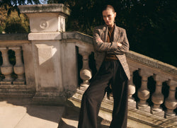 A.P.C. x Anastasia Barbieri campaign image, featuring a female model wearing a blazer, brown sweater, and dark pants, on a stair case