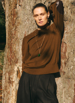 A.P.C. x Anastasia Barbieri campaign image, featuring a female model wearing a necklace, a brown sweater, and dark pants, by a tree.