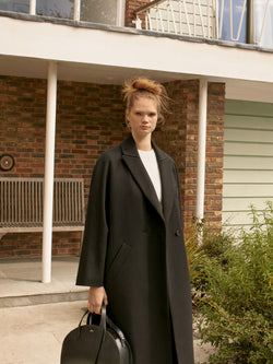 A.P.C. The Brand - woman wearing black coat and holding a bag standing in front of a house.