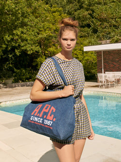 A.P.C. The Brand - woman wearing plaid romper and holding a tote bag standing in front of a pool.